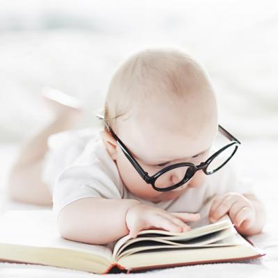 A baby in a white onesie lays on top of a book, grabbing the edge of a chunk of pages in one hand and laying the other flat on the book as if reading it. On top of their serious face are perched a pair of precarious, oversized black spectacles.