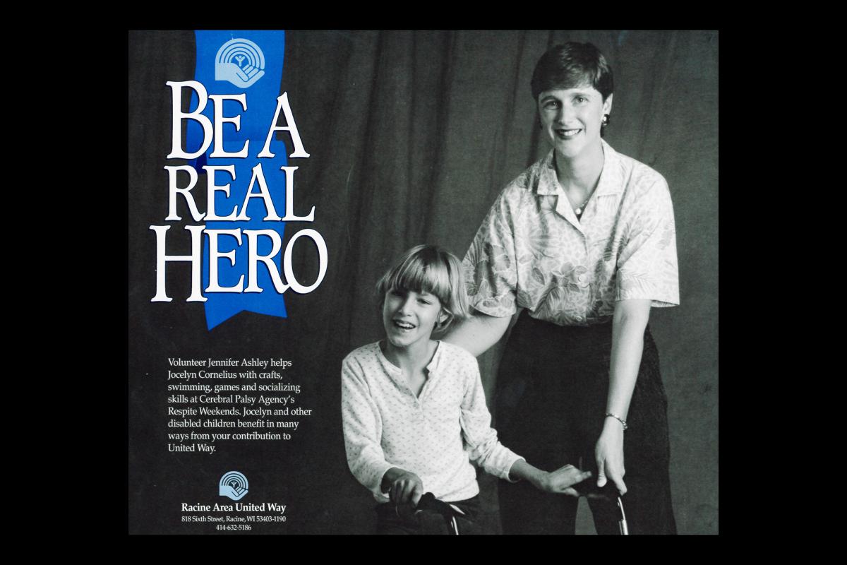 A poster from United Way's days as Racine Area United Way. The background is a black and white photo of a young adult with dark, buzzed hair, lipstick and earrings putting their hand on the back of a child beside them, who has a blonde bowl cut and stands with a walker. Both are smiling. Beside them, a blue ribbon with an early version of the United Way logo drops down and says, "Be a real hero." Below is more text that says, "Volunteer Jennifer Ashley helps Jocelyn Cornelius with crafts, swimming, games and socializing skills at Cerebral Palsy Agency's Respite Weekends. Jocelyn and other disabled children benefit in many ways from your contribution to United Way. Racine Area United Way. 818 Sixth Street, Racine, WI 53403-1190. 414-632-5186."