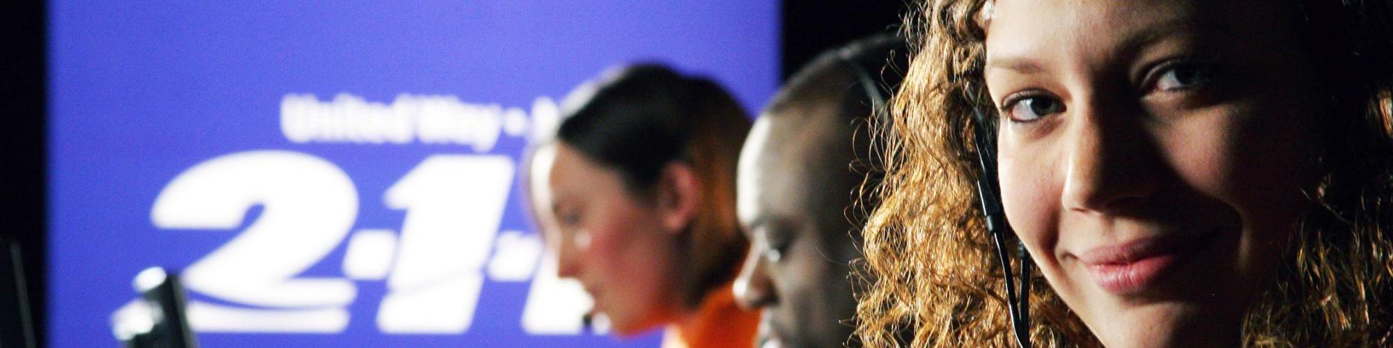 Three call center workers wear earpieces on their ears, gazing at their computers. Behind them, a blue screen with the 2-1-1 logo on it shines. The closest of the workers looks up at the camera with a knowing smile.