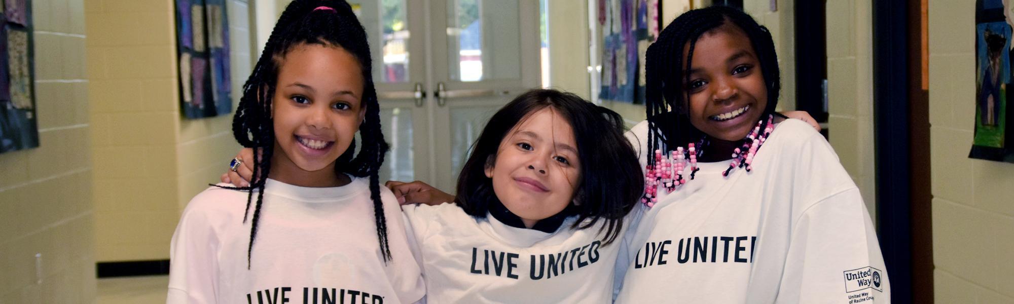 Three kids in the hall of Knapp Elementary wearing oversized Live United shirts wrap their arms around each other's shoulders and strike cool poses for the camera.